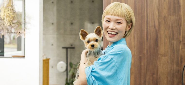 ペットサロンのオーナーがお客様のペットを抱いて、ペットサロンの前で笑顔でこちらを向いている様子の写真